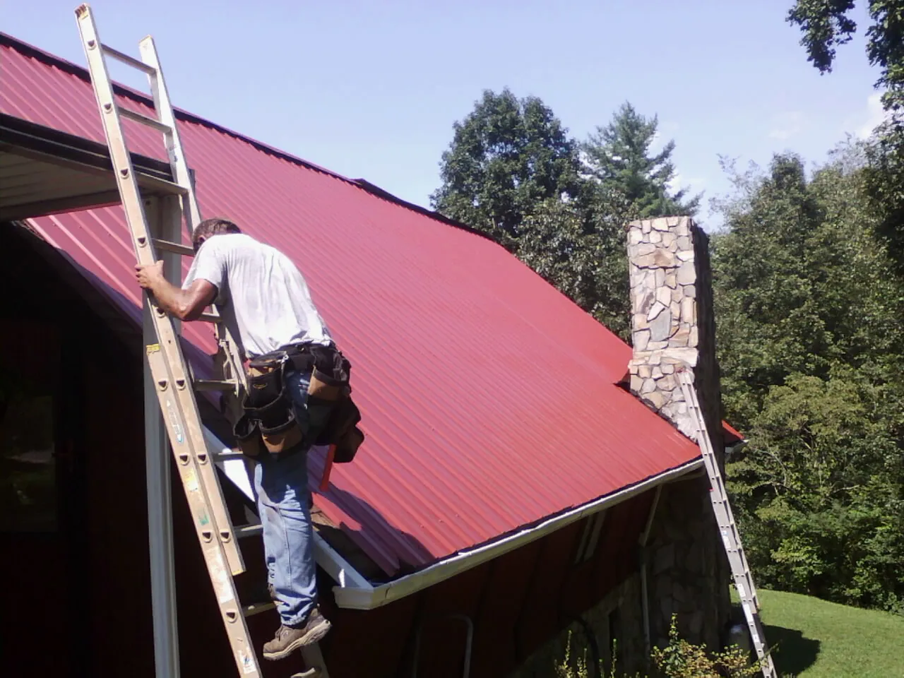 Roofer on a ladder beside a red metal roof and stone chimney — Soffit & Fascia Repair by Roofing Brothers in Windsor Mill